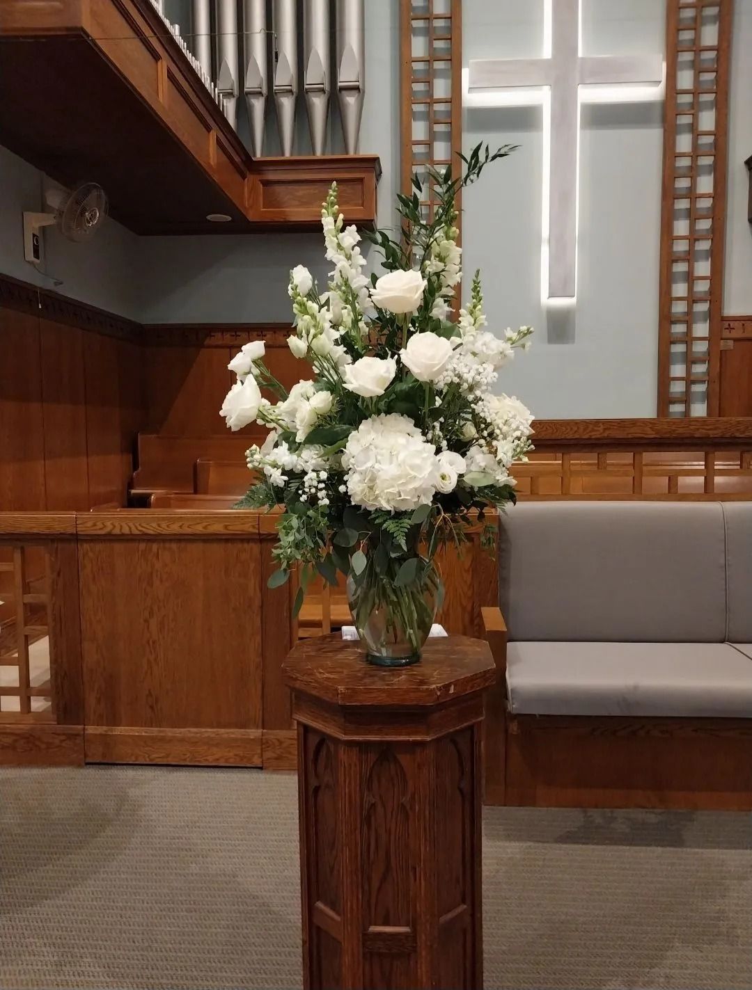 Floral arrangement of white flowers in a vase on a wooden pedestal inside a church.