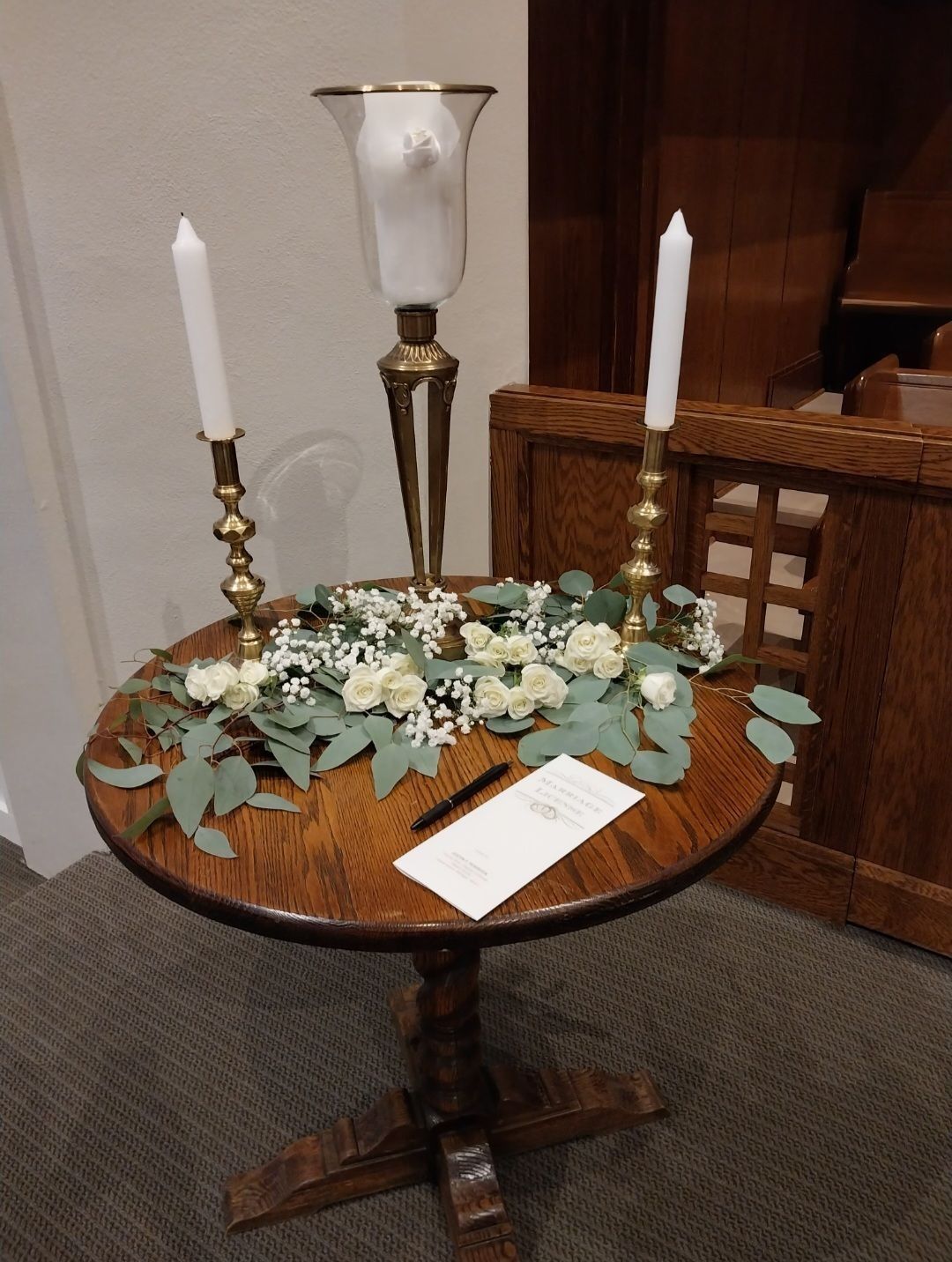 A decorated round wooden table in a church with candles, flowers, and a sign.