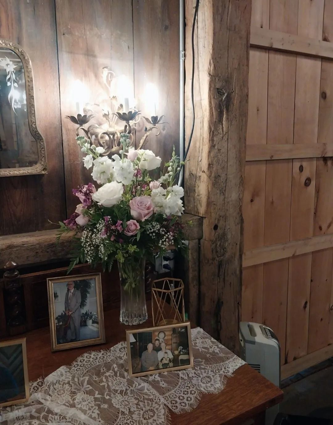Floral arrangement with framed photos on a wooden table, lit by sconce. Barn setting.