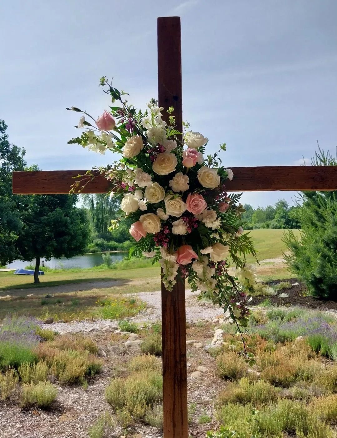 Wooden cross adorned with a floral arrangement of white and pink roses, set outdoors with a landscape background.