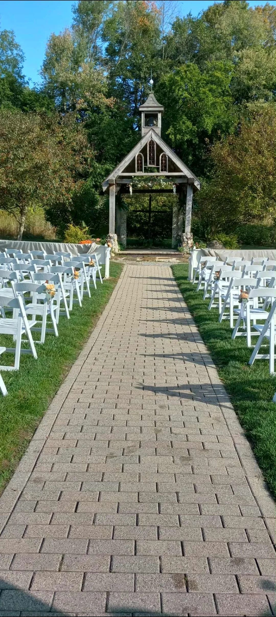 Stone path leads to a wooden structure with rows of white chairs on either side; trees in the background.