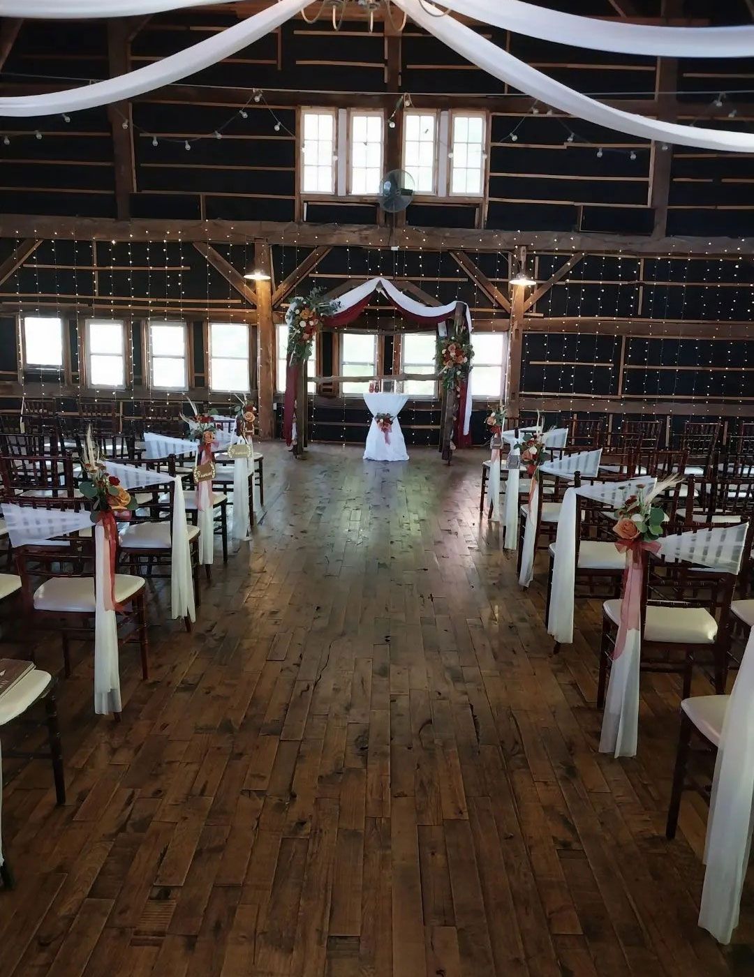 Wedding ceremony inside a barn. Wooden floor, chairs with white fabric. Ceremony arch decorated with flowers and fabric.