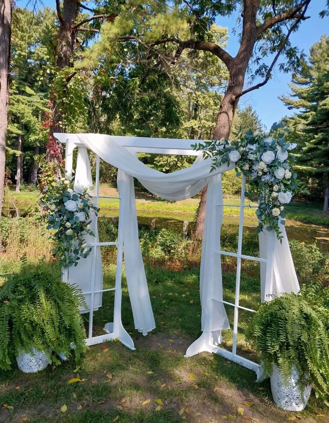 White wedding arch with draping fabric and floral arrangements, in a park setting.