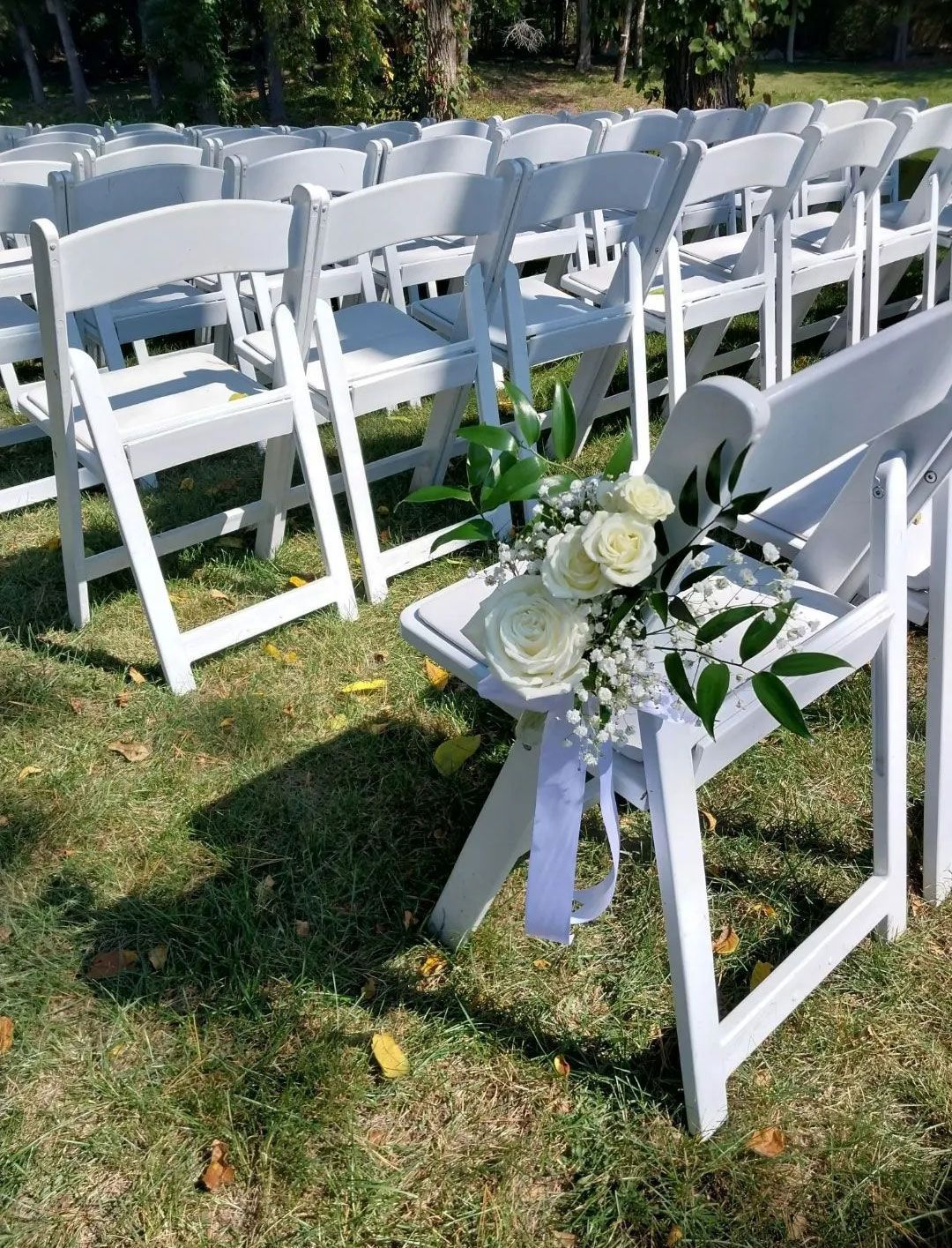 White chairs set up for an outdoor wedding ceremony, with floral decoration on the front chair.