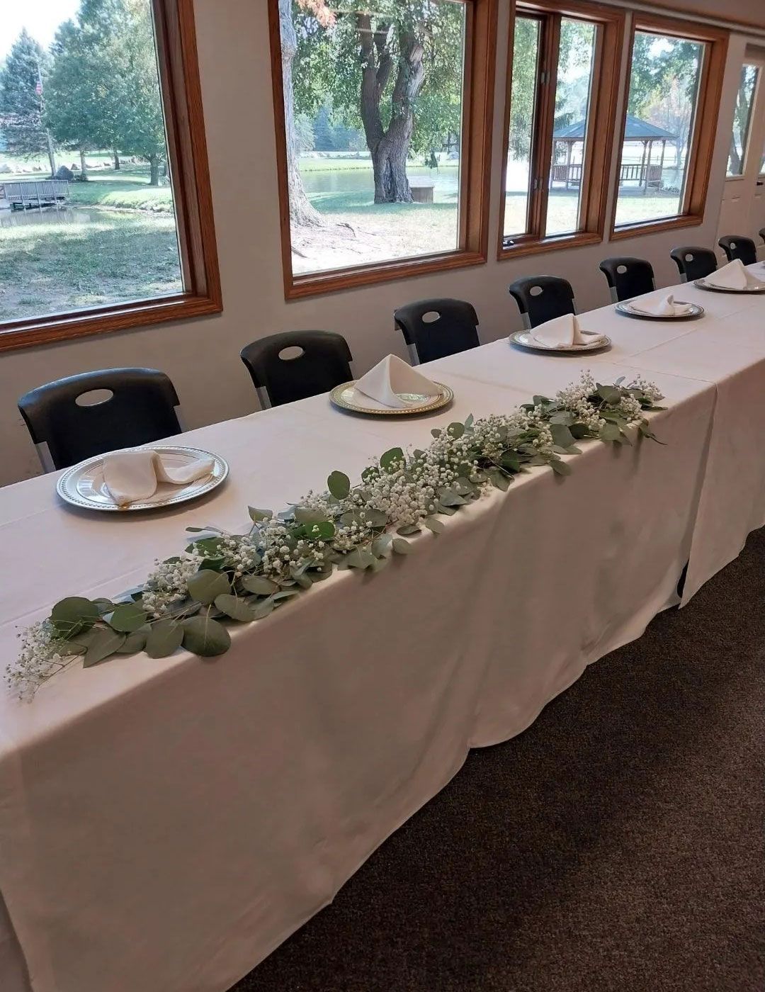 Long banquet table set with white tablecloth, place settings, and greenery garland. Windows with outdoor view.