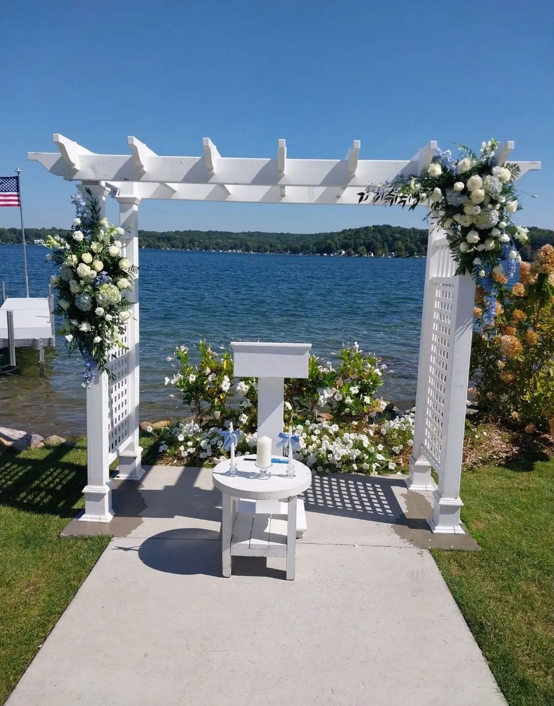 Wedding ceremony setup by a lake with floral arrangements. White arbor, podium, and table with blue water and sky backdrop.