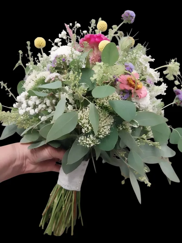 Hand holding a bouquet of mixed wildflowers in shades of pink, white, yellow, and purple with green eucalyptus leaves.