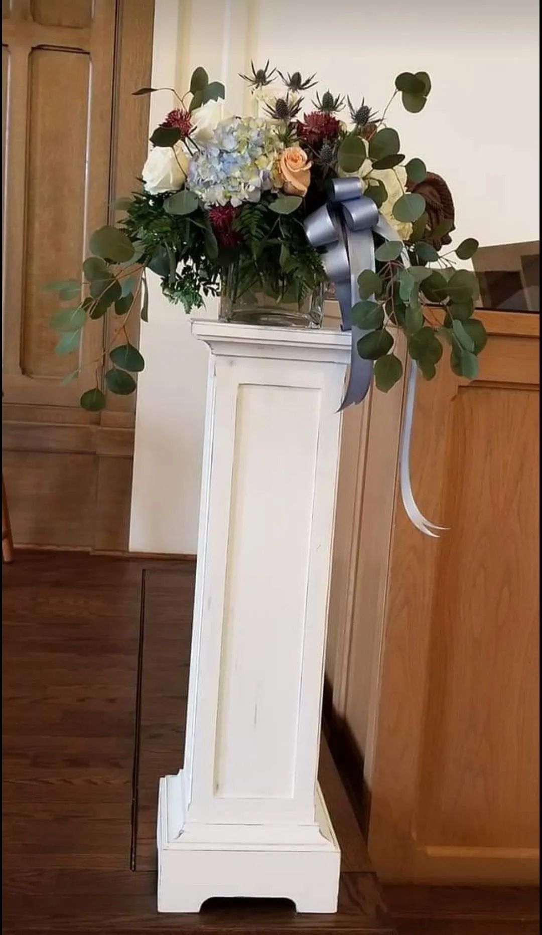Floral arrangement on a white pedestal, with blue and peach flowers, silver ribbon, inside a wood-paneled room.