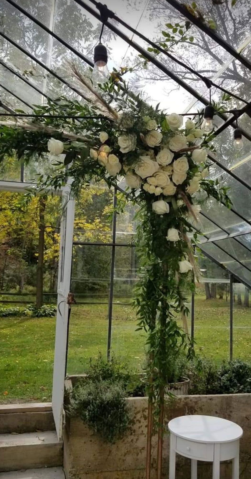 Floral archway with white roses and greenery in a glass greenhouse setting.
