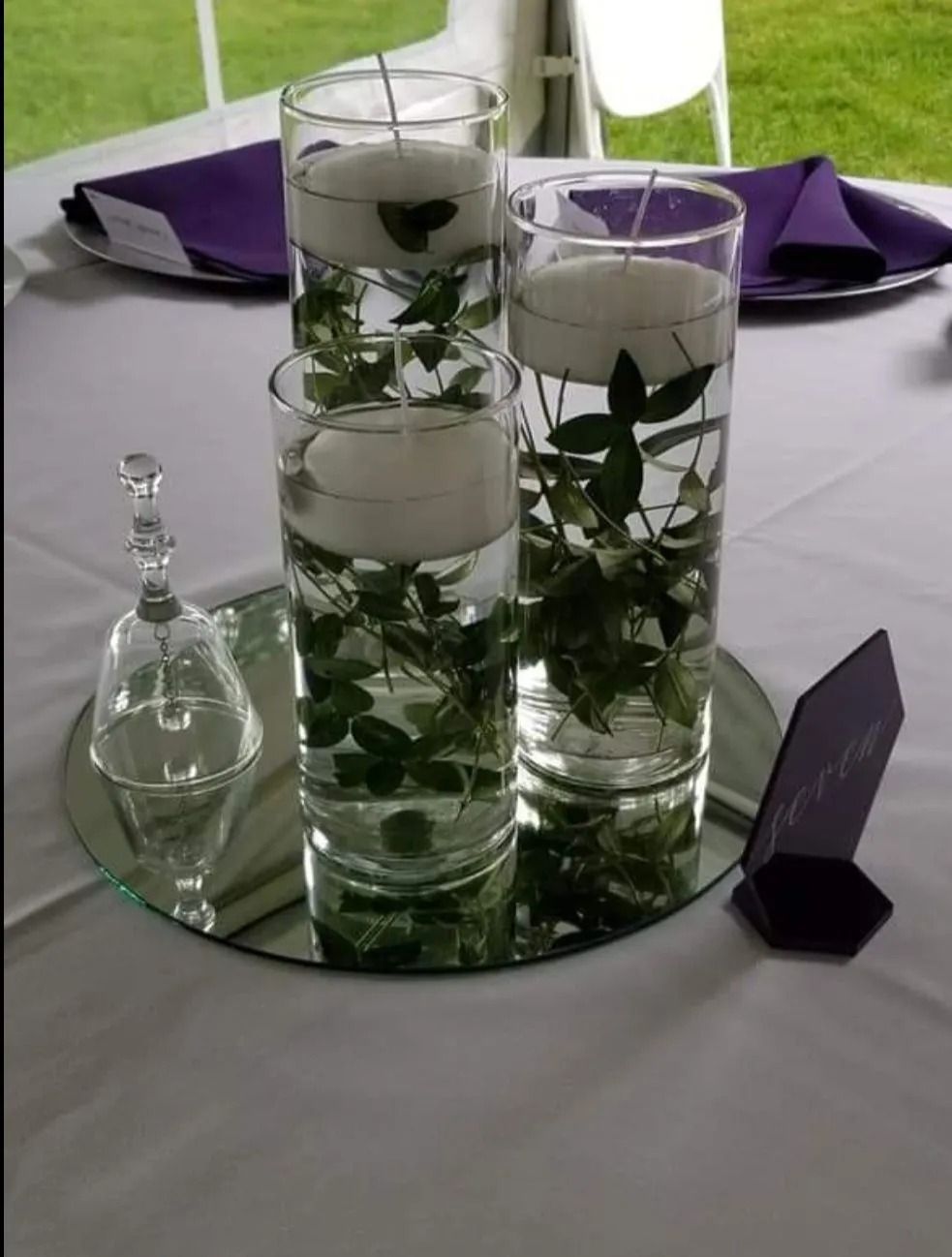 Centerpiece with floating candles and greenery in glass vases on a mirrored tray; purple napkins.