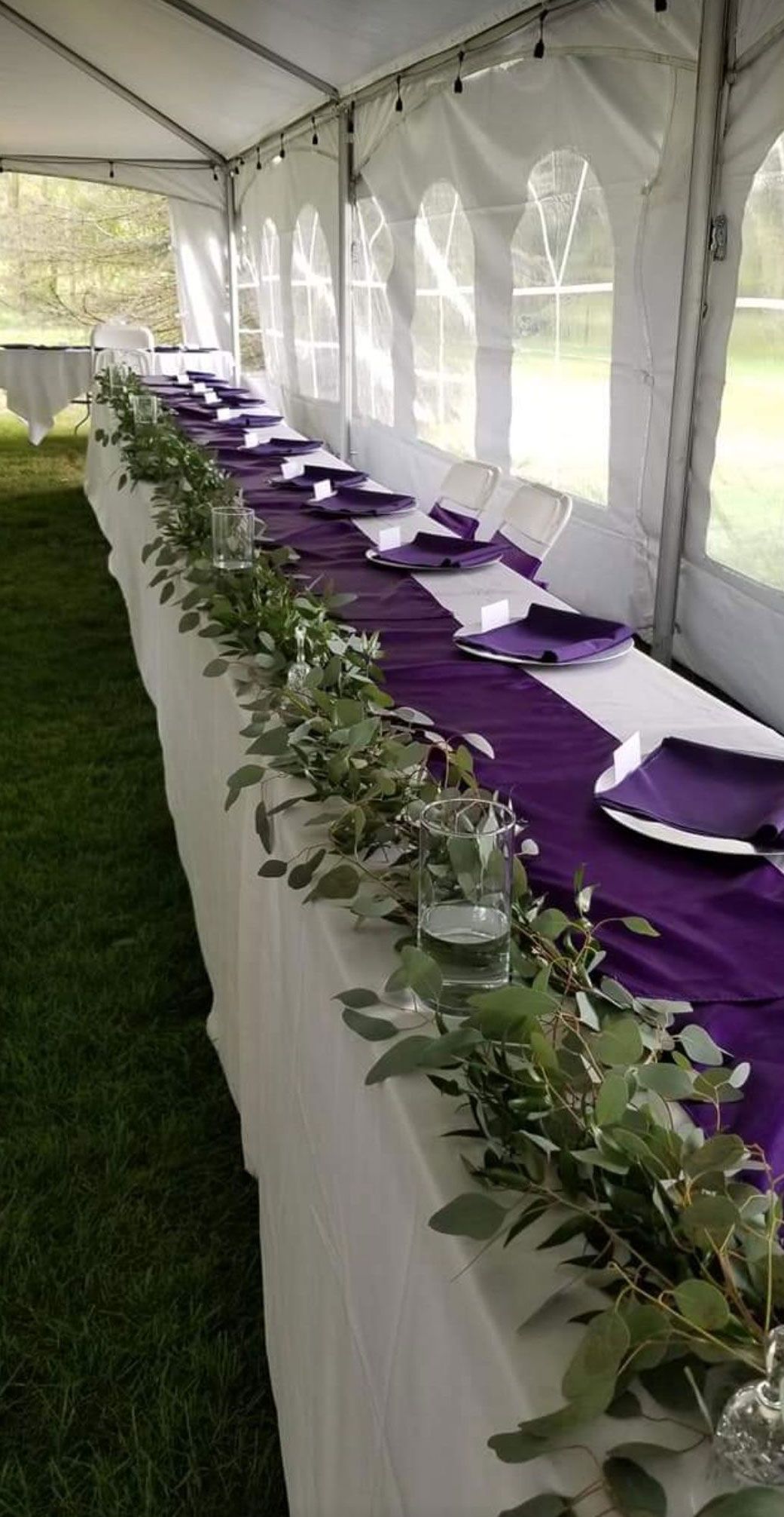 Long table set for a formal event, white tablecloth, purple runner, greenery garland, white plates, purple napkins, tent setting.