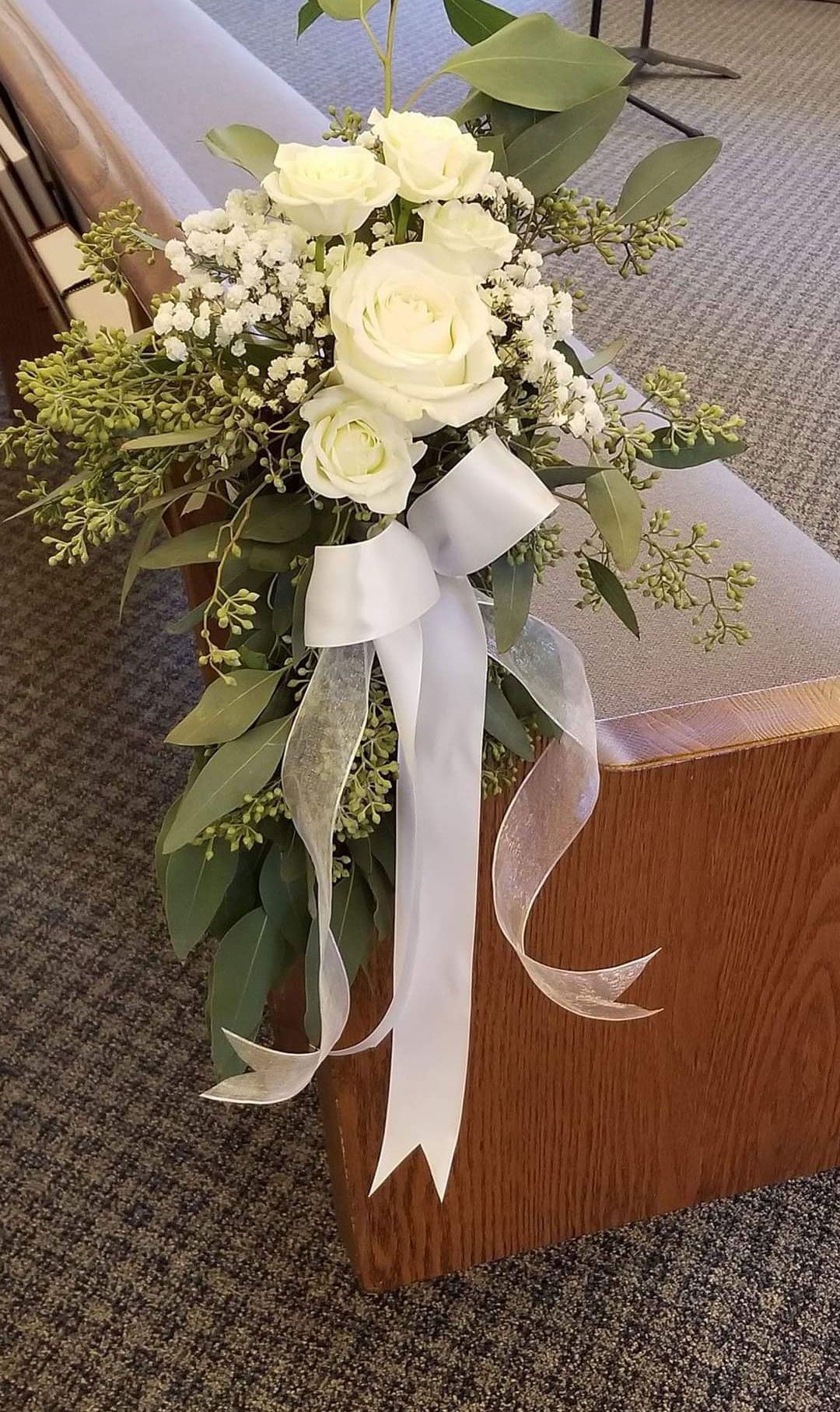 White rose floral arrangement with ribbon tied to a church pew.