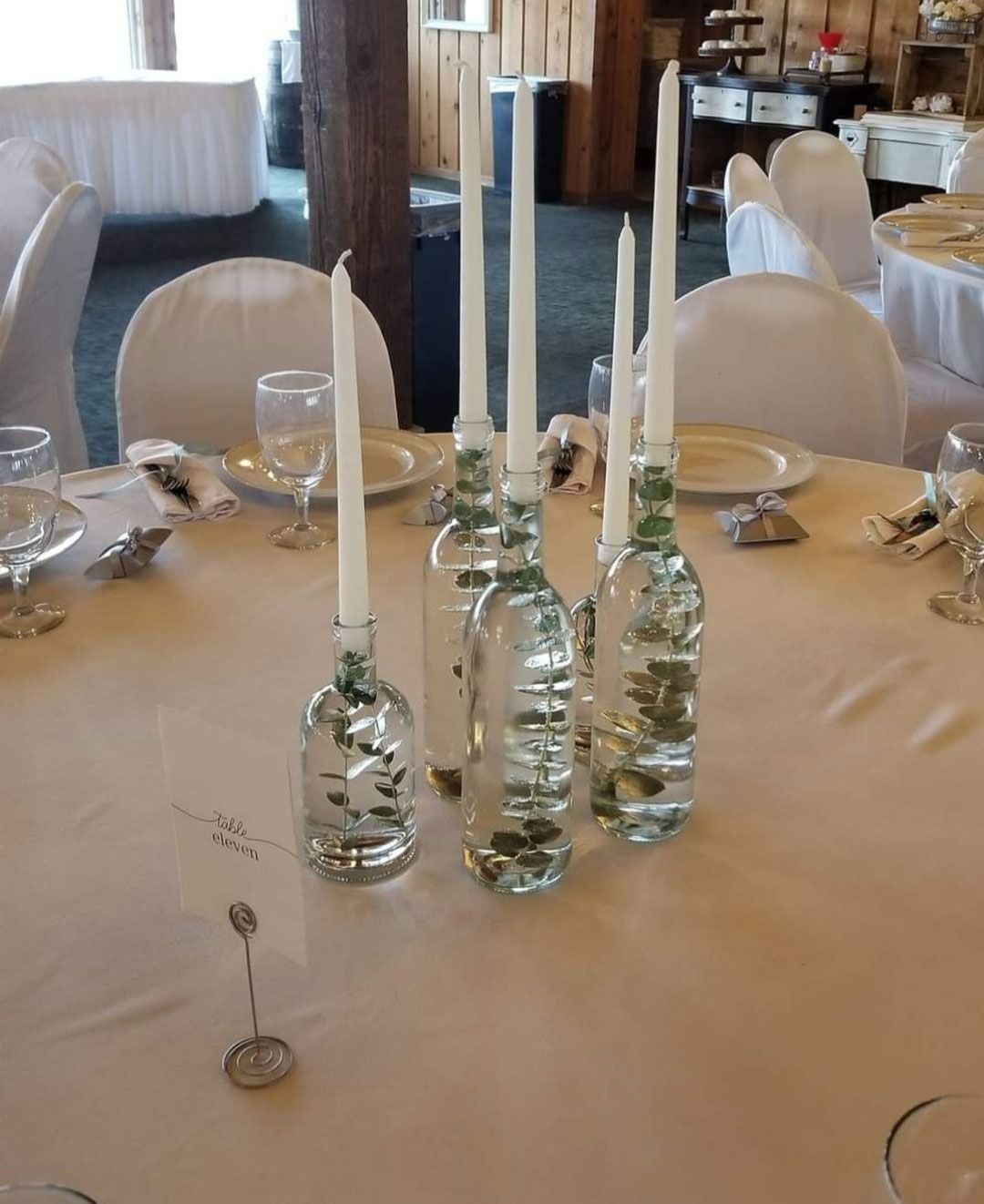 Centerpiece of tall white candles in water-filled glass bottles on a table set for a wedding.