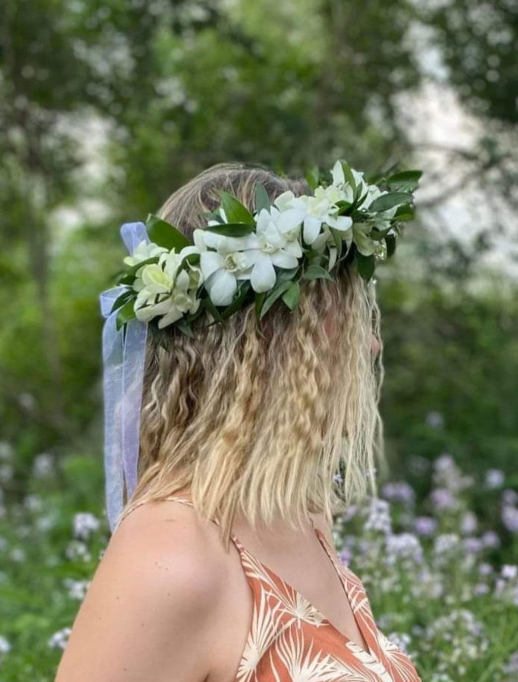 Woman with curly blonde hair wearing a white flower crown, standing outdoors in a garden setting.