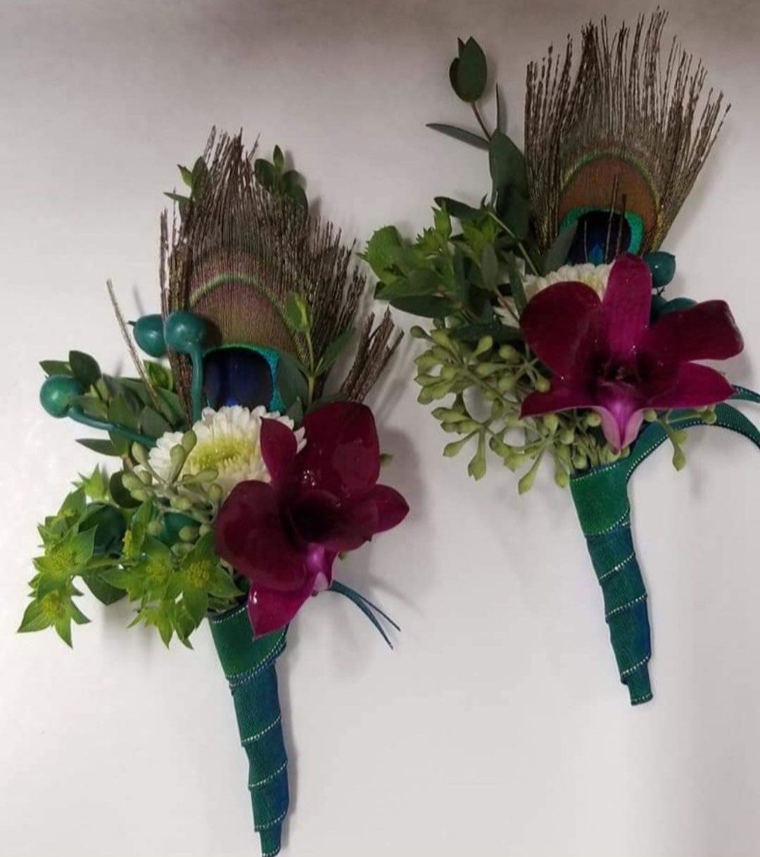 Two peacock feather and orchid boutonnieres wrapped in green tape, on a white surface.