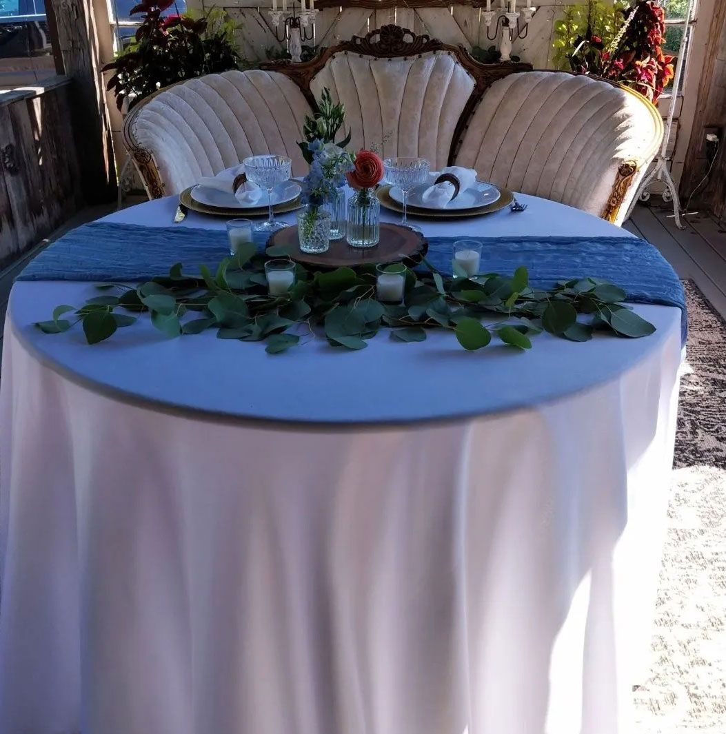 Round table set for two with blue runner, foliage, and ornate sofa backdrop.