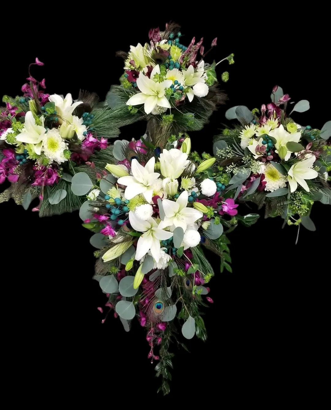 Floral arrangement in the shape of a cross, white lilies, purple accents, green foliage, black background.
