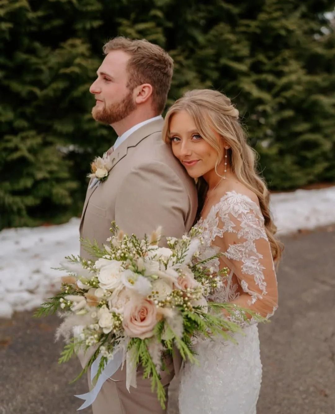 Bride in lace dress leans on groom's back, holding bouquet. They stand outside near green trees.