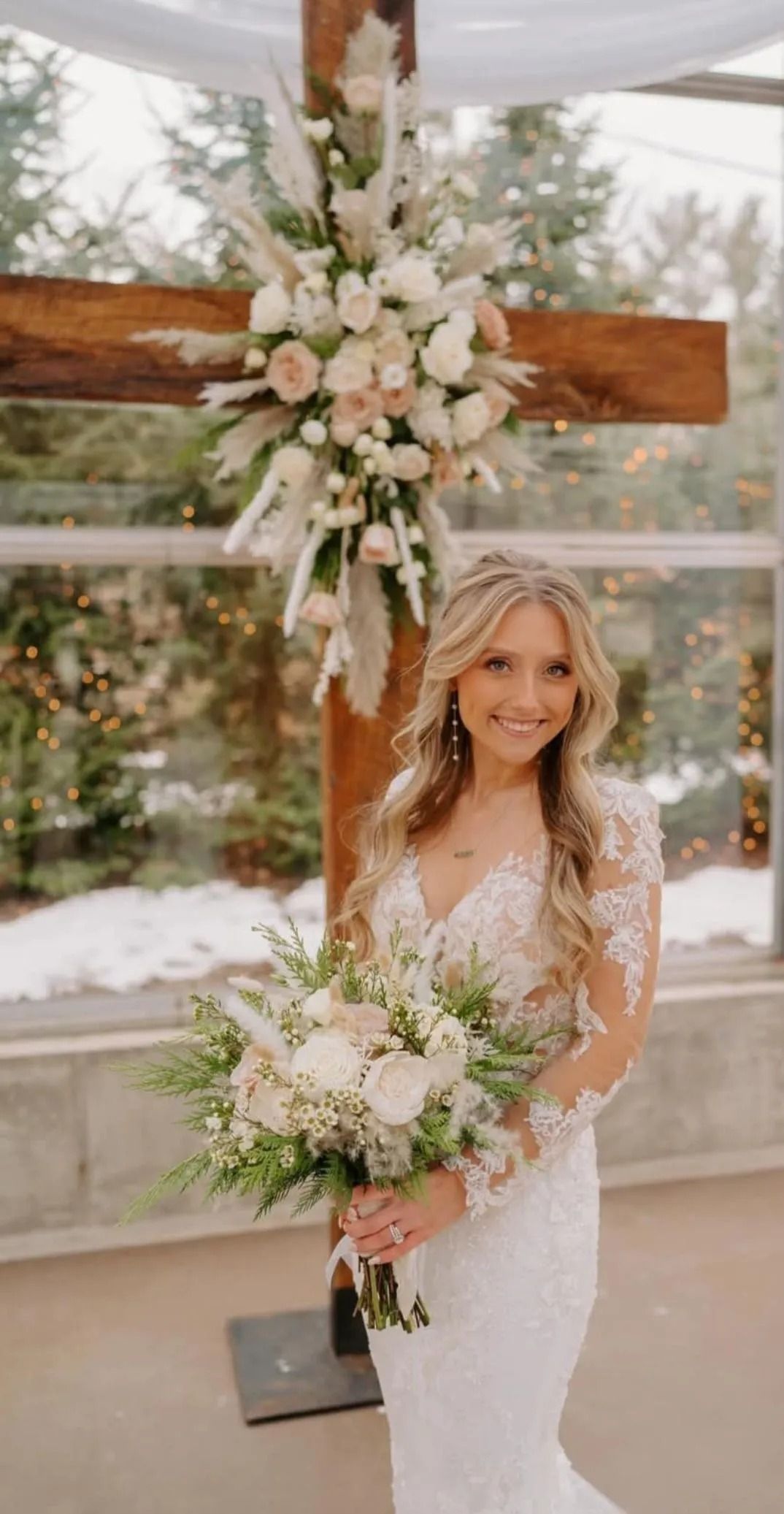 Bride in lace dress holding bouquet, smiling in front of a wooden cross decorated with flowers.