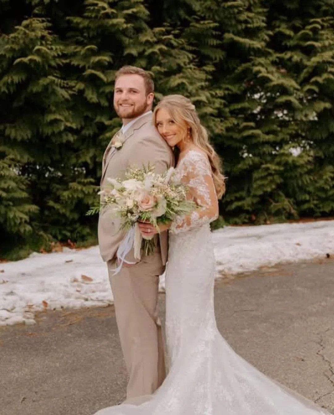 Couple in wedding attire: beige suit, lace gown, holding bouquet, posing outdoors near snow.