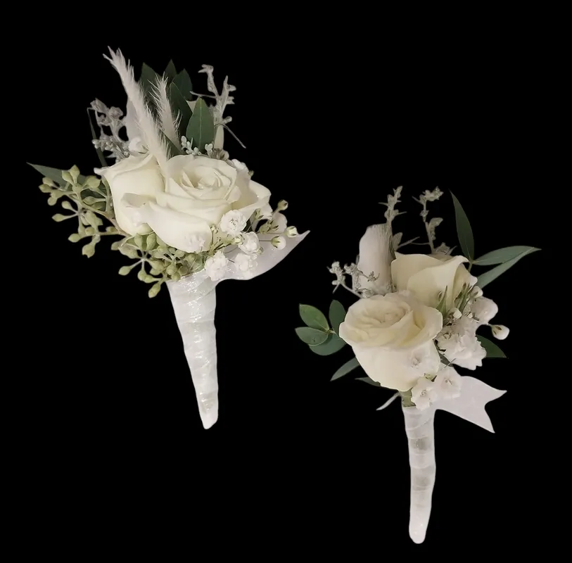 Two white rose boutonnieres with greenery and ribbon on a black background.
