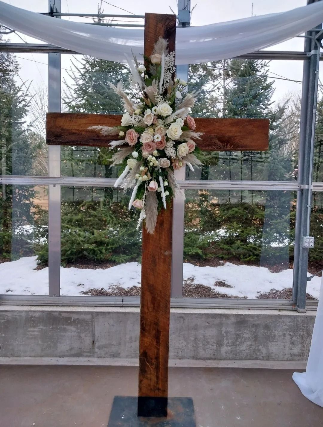 Wooden cross decorated with flowers, in a greenhouse setting with trees visible outside.