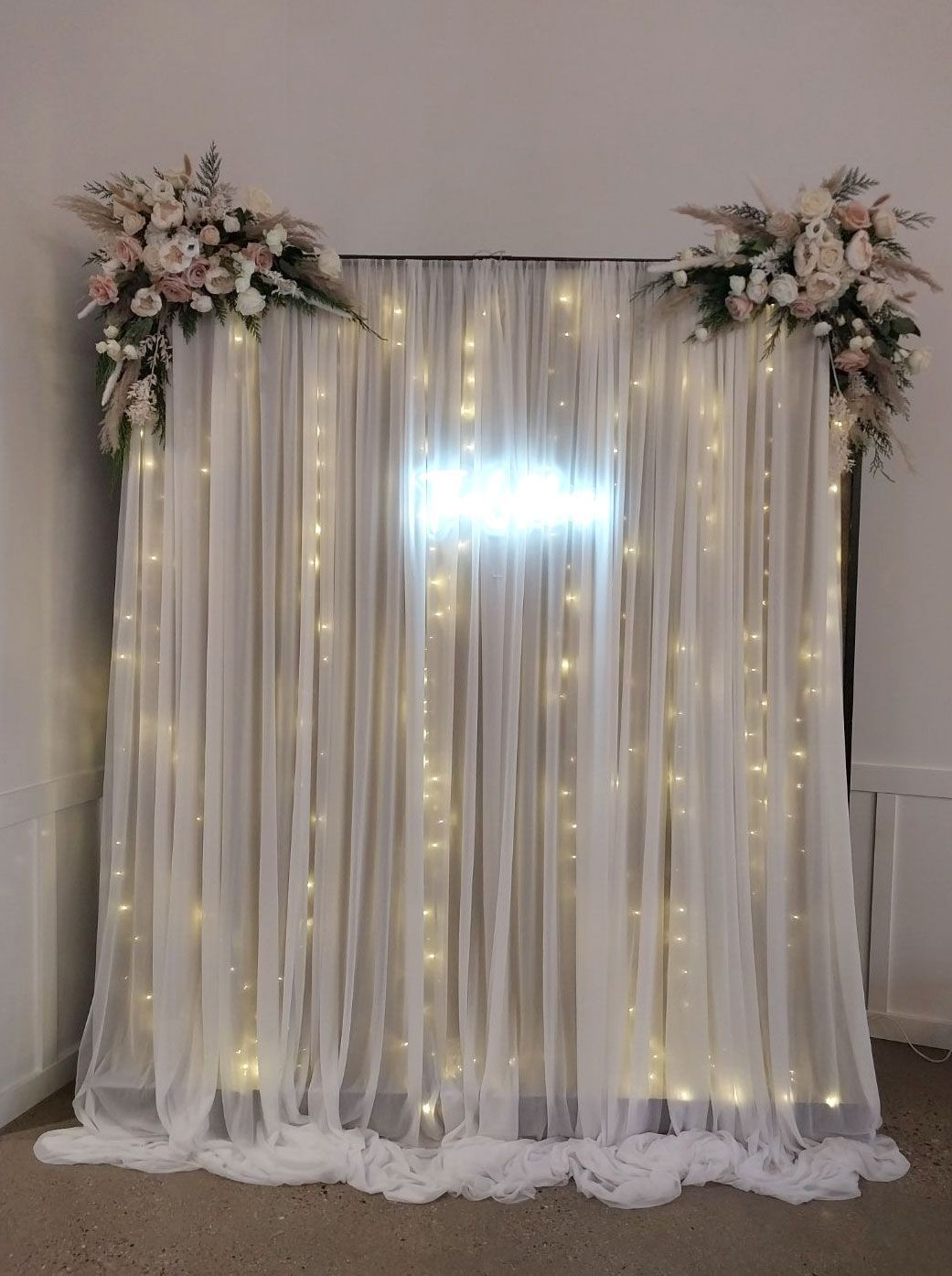 Sheer white curtain backdrop with string lights, floral arrangements, and a glowing sign.