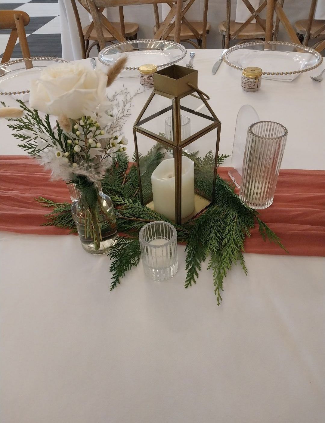 Centerpiece on white tablecloth: lantern, candles, flowers, greenery, and orange runner.