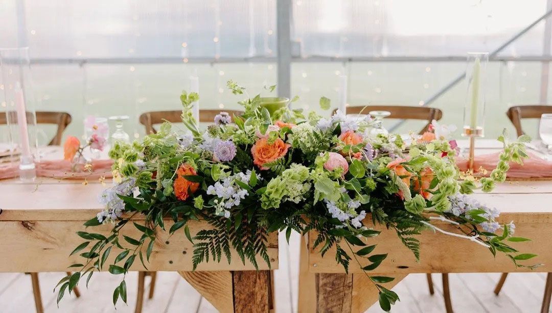 Wooden table with floral centerpiece, pastel colors, inside a greenhouse.