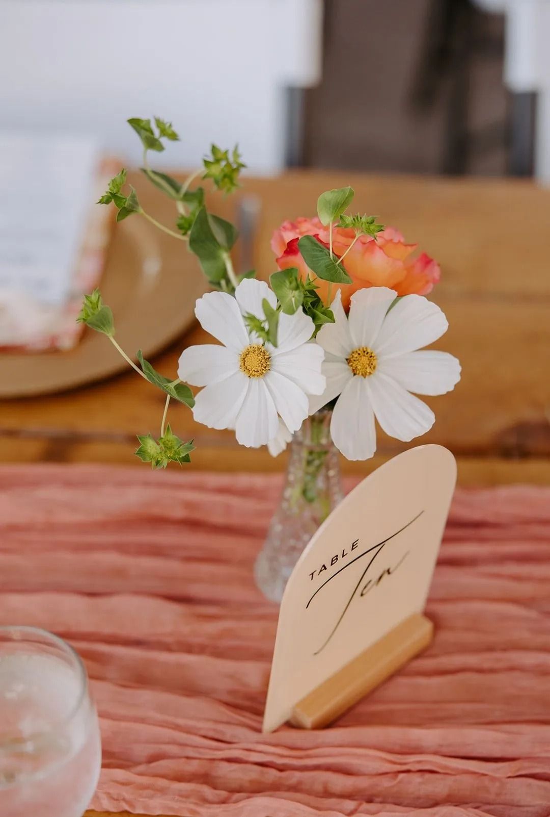 White and orange flowers in a vase on a pink table runner next to a wooden table number.
