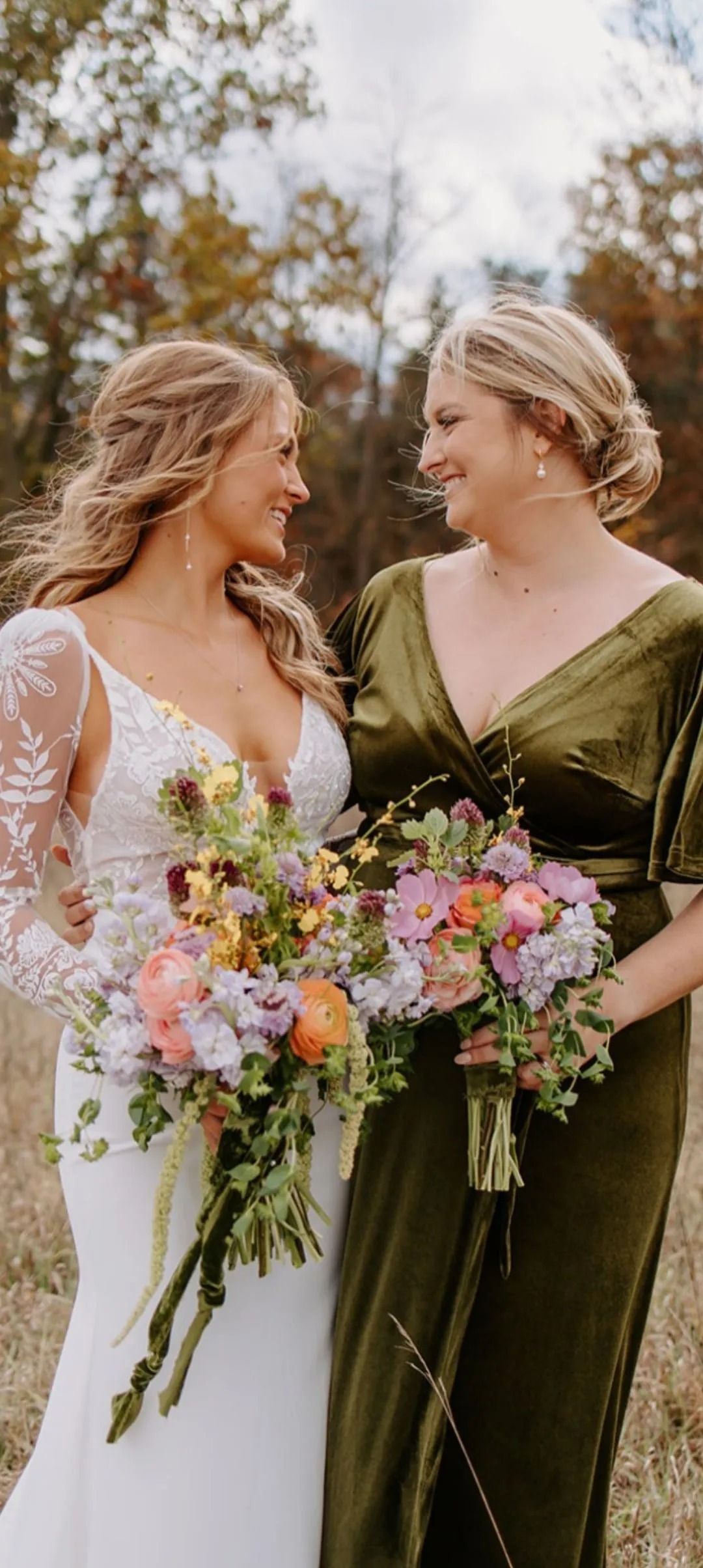 Two women smiling, holding bouquets. One in a white dress, the other in a green velvet dress. Outdoors.