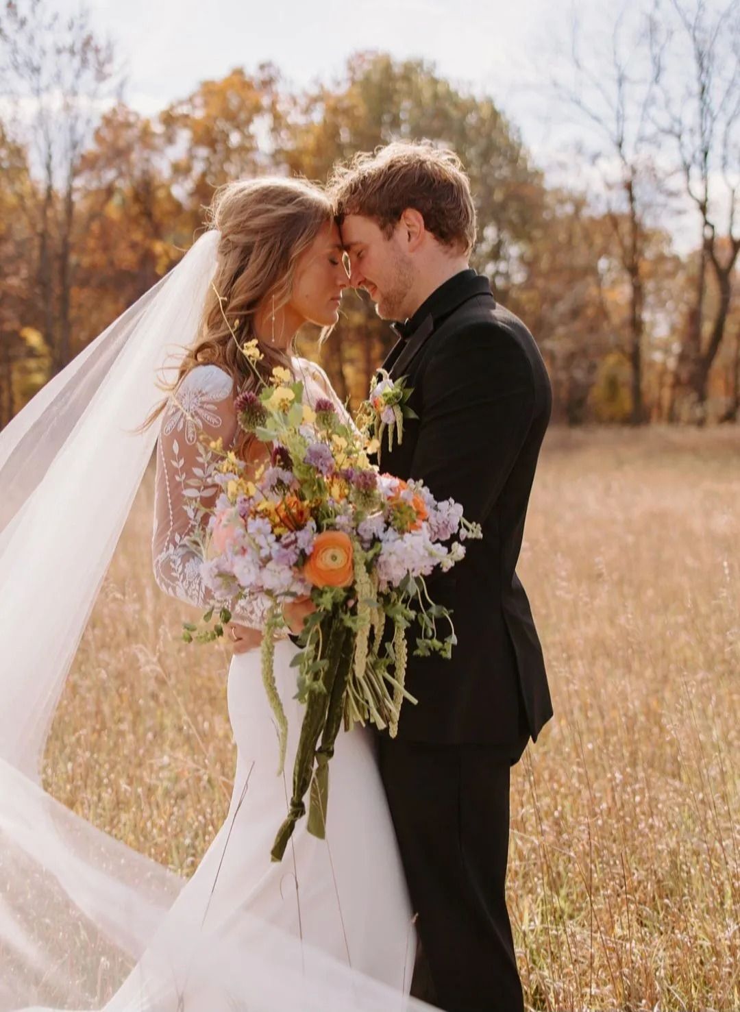Bride and groom embrace in a field, woman in white dress holding bouquet, autumn foliage backdrop.