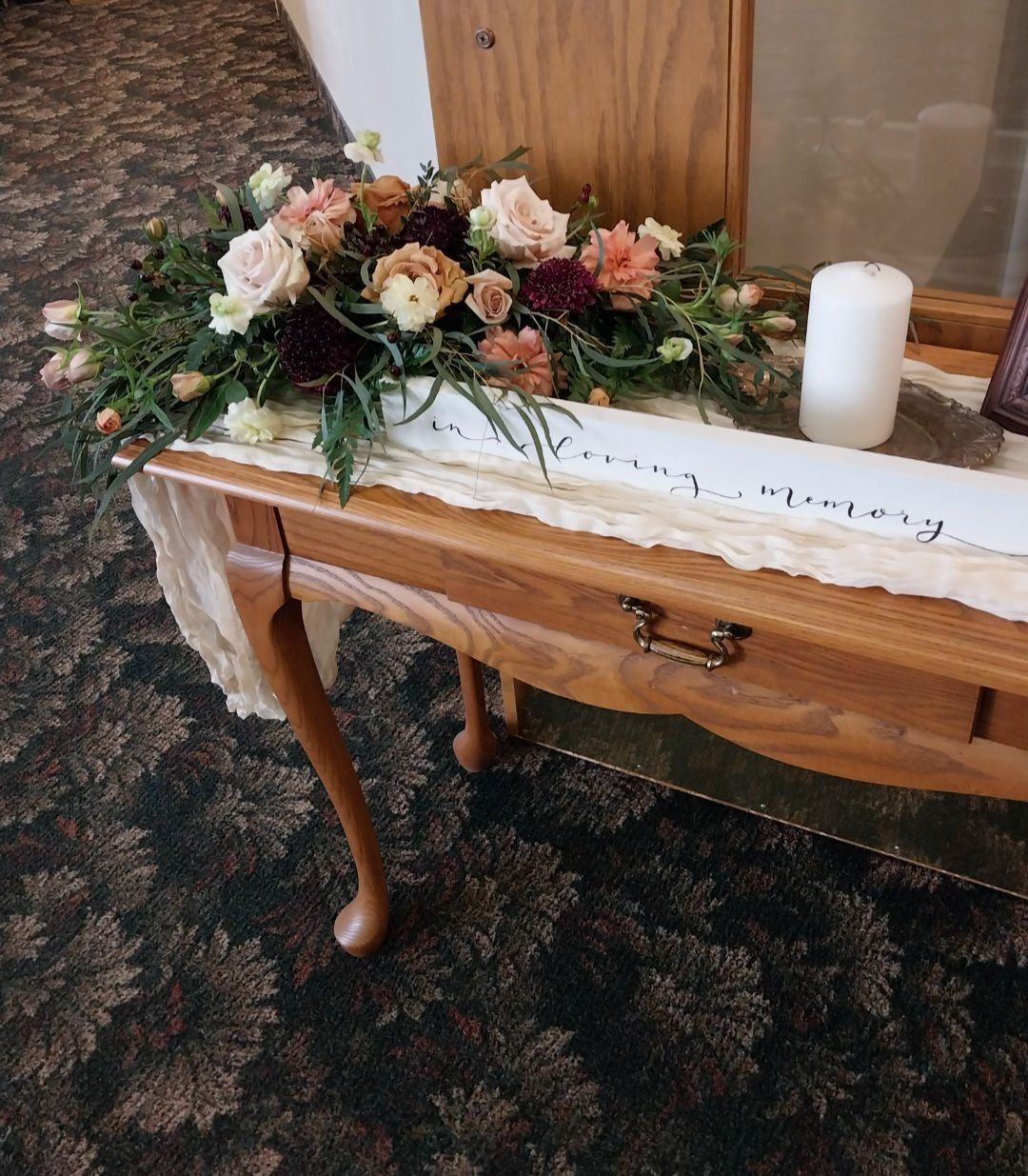 Floral arrangement on a wooden table, with a candle and banner on the right, set on patterned carpet.