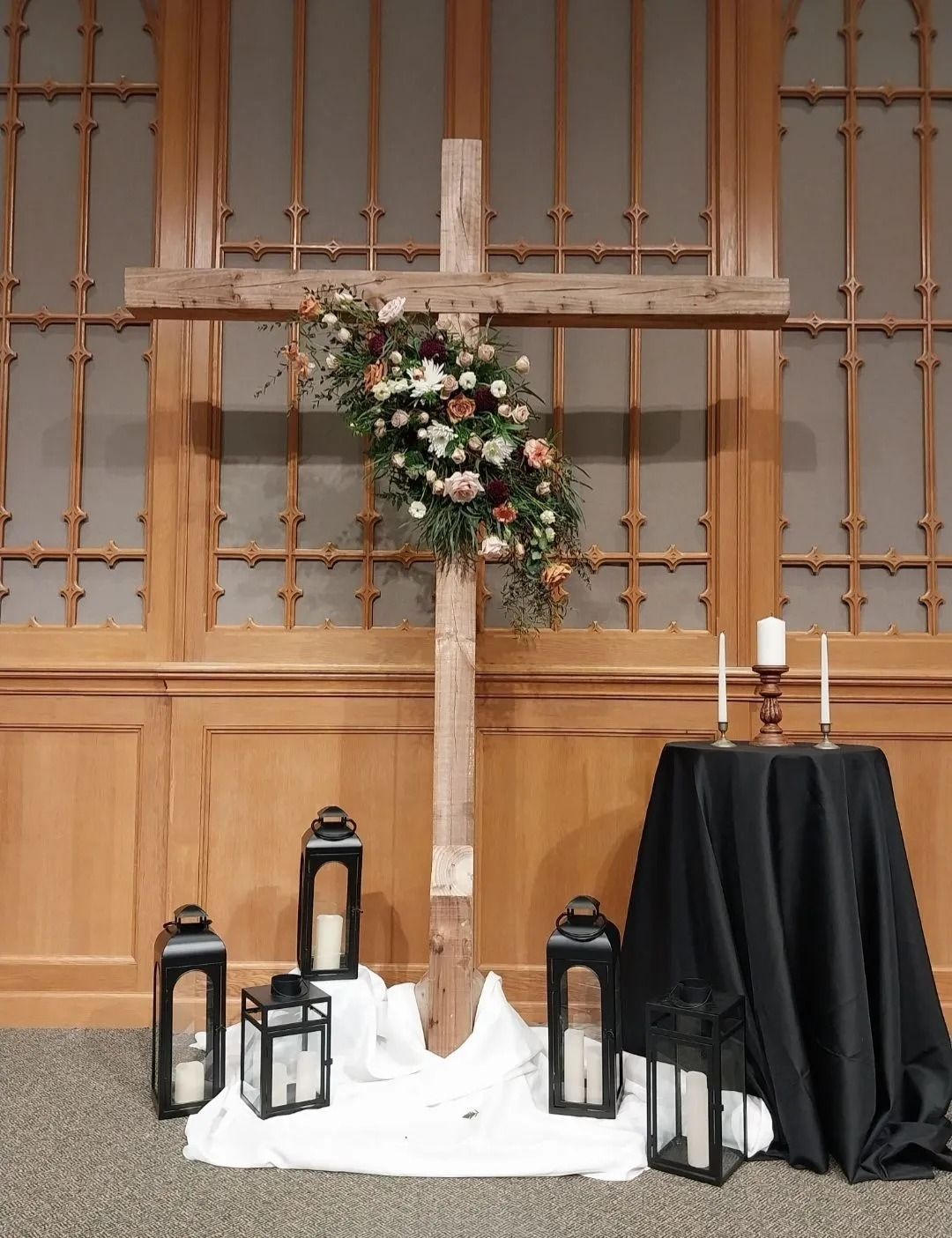 Wooden cross with floral arrangement, candles, and lanterns in front of a wood-paneled wall.