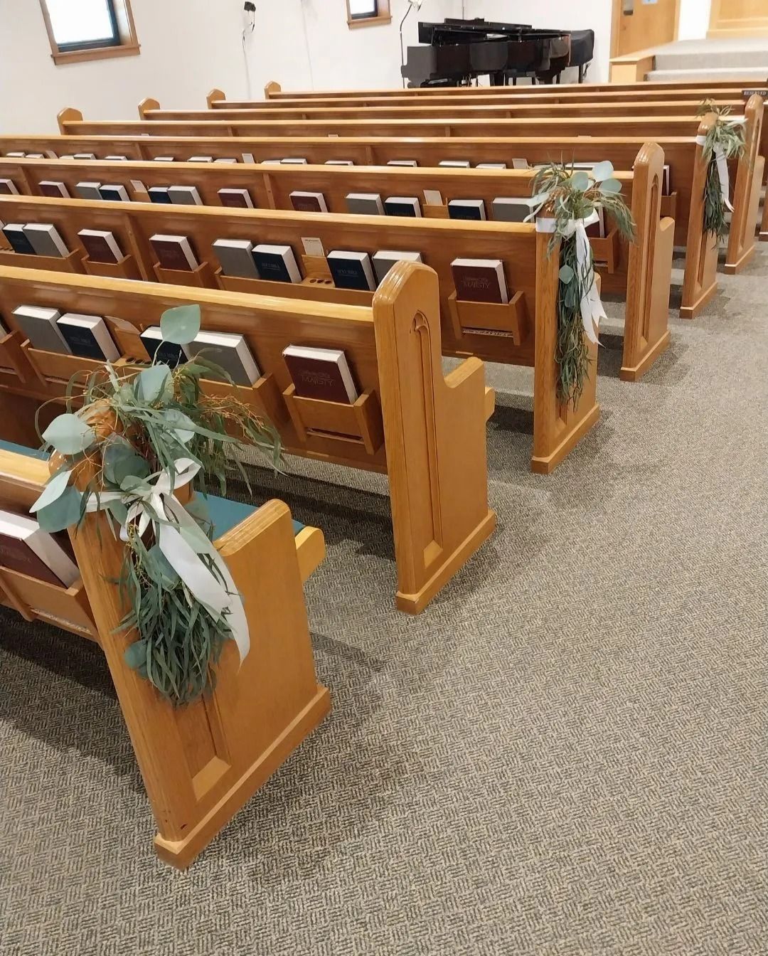 Rows of wooden church pews decorated with greenery and white ribbons. Carpeted floor, piano in the background.