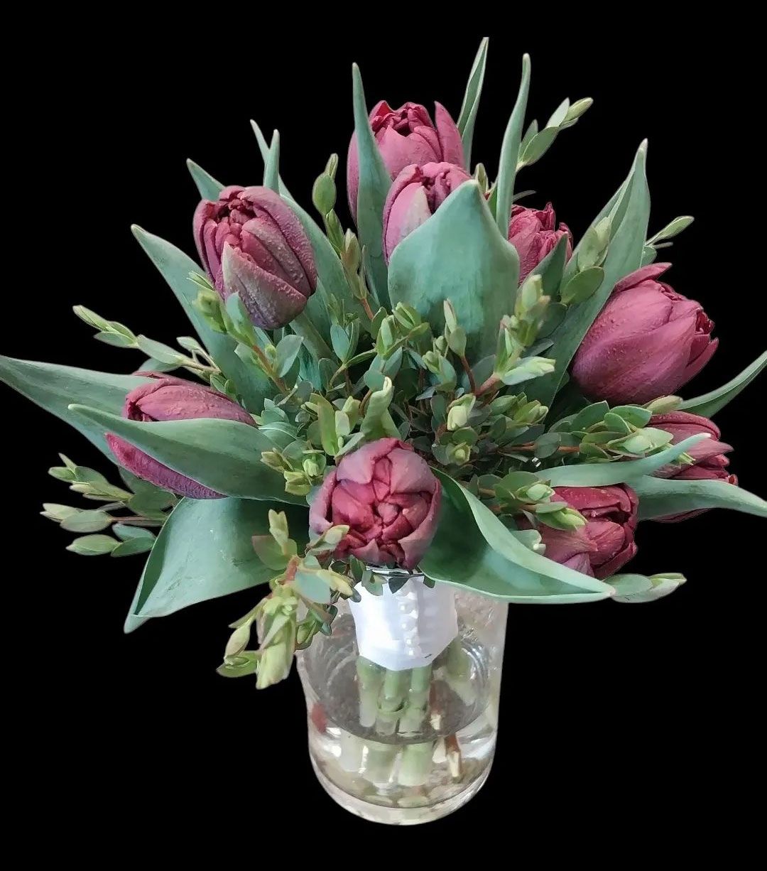 Bouquet of dark red tulips in a glass vase, surrounded by green leaves and small green sprigs, set against a black background.