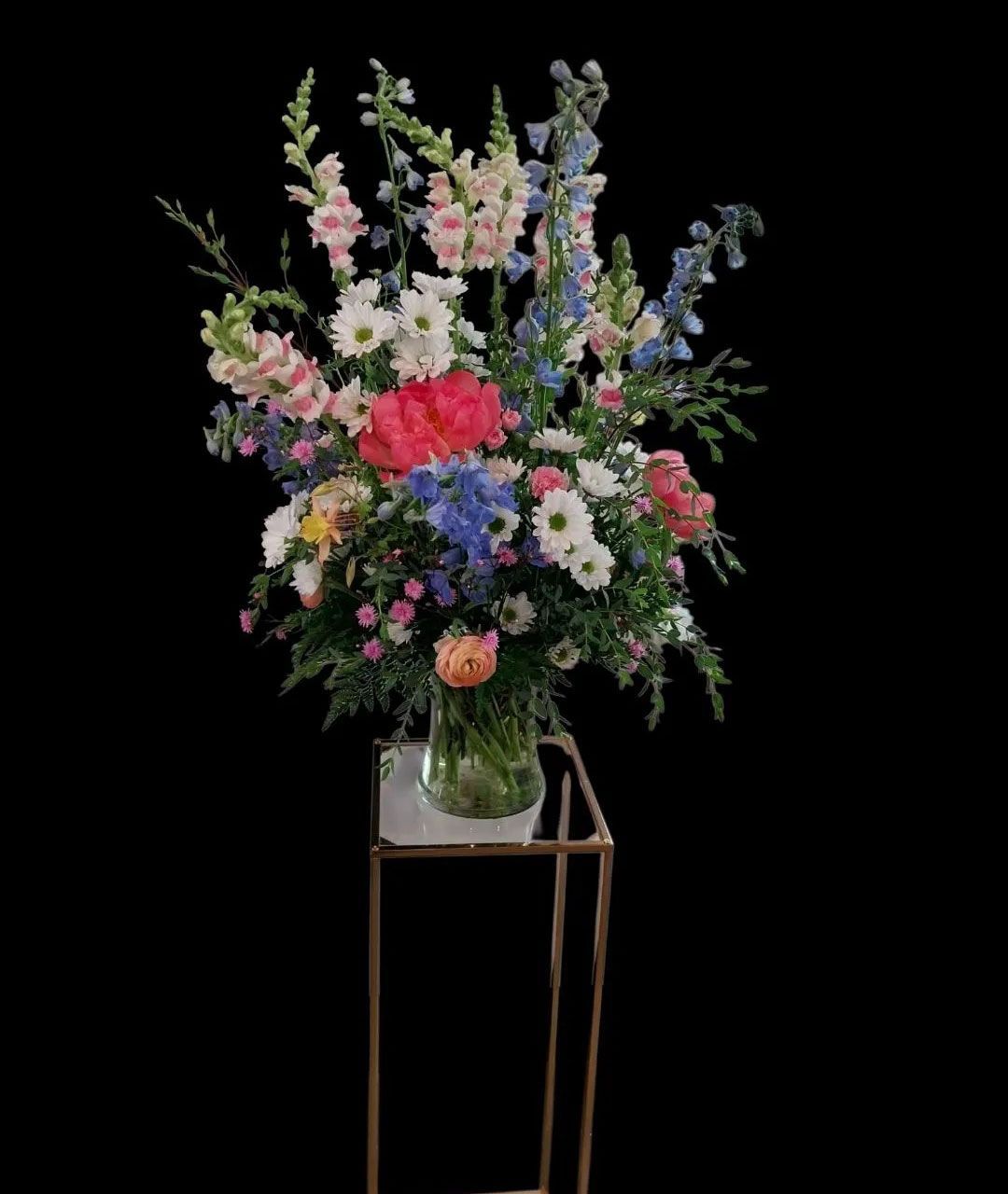 Colorful floral arrangement in glass vase on a gold stand against a black background.