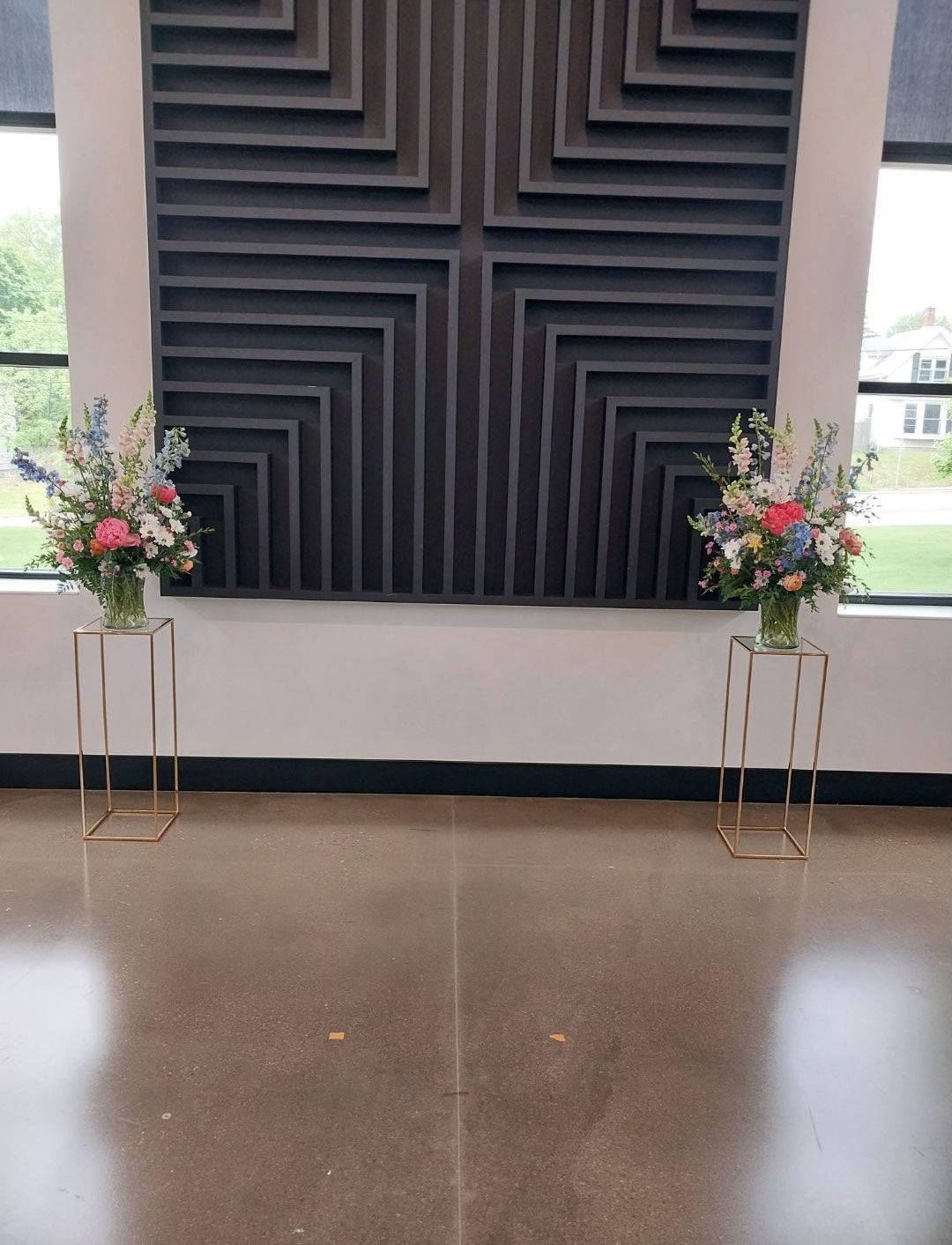 Two floral arrangements on gold stands flank a dark geometric wall art in a modern space.