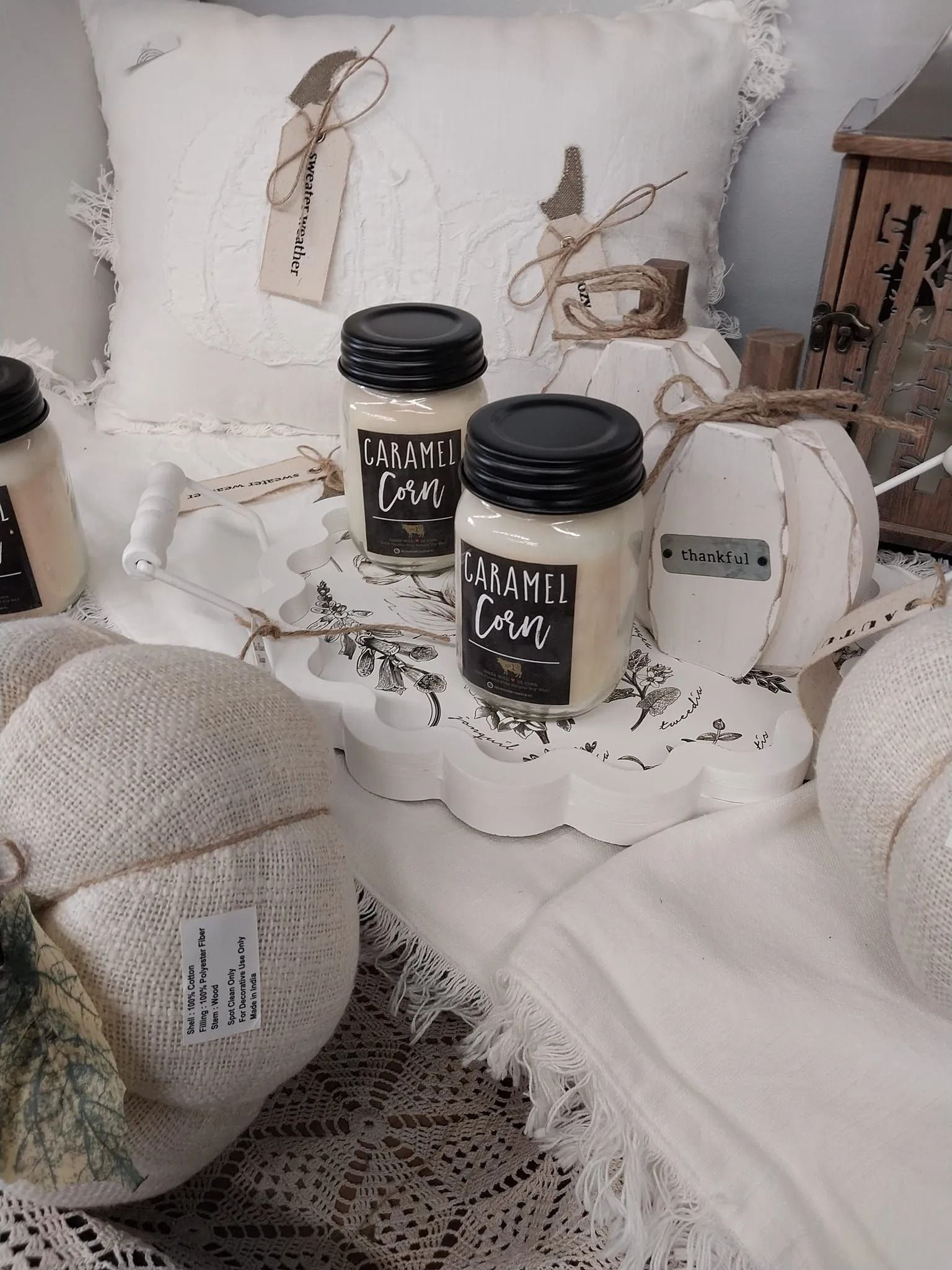 Fall-themed display: cream-colored pumpkins, caramel corn candles, decorative pillow, white and lace accents.