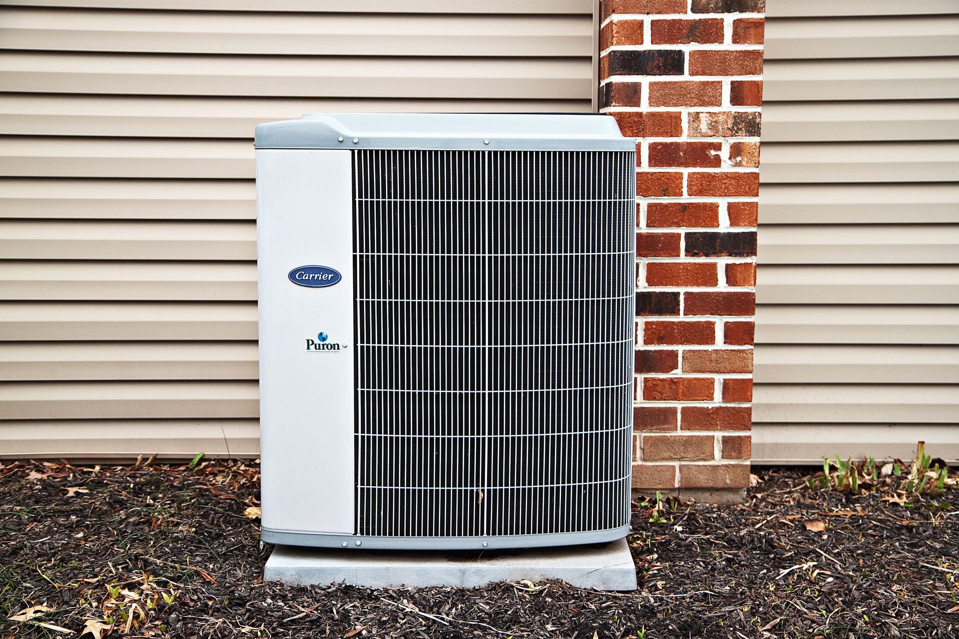 Air conditioning unit outside a building, set on a concrete base next to brick column and horizontal siding.