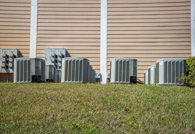 Air conditioning units lined up on grass against a building with light brown siding.