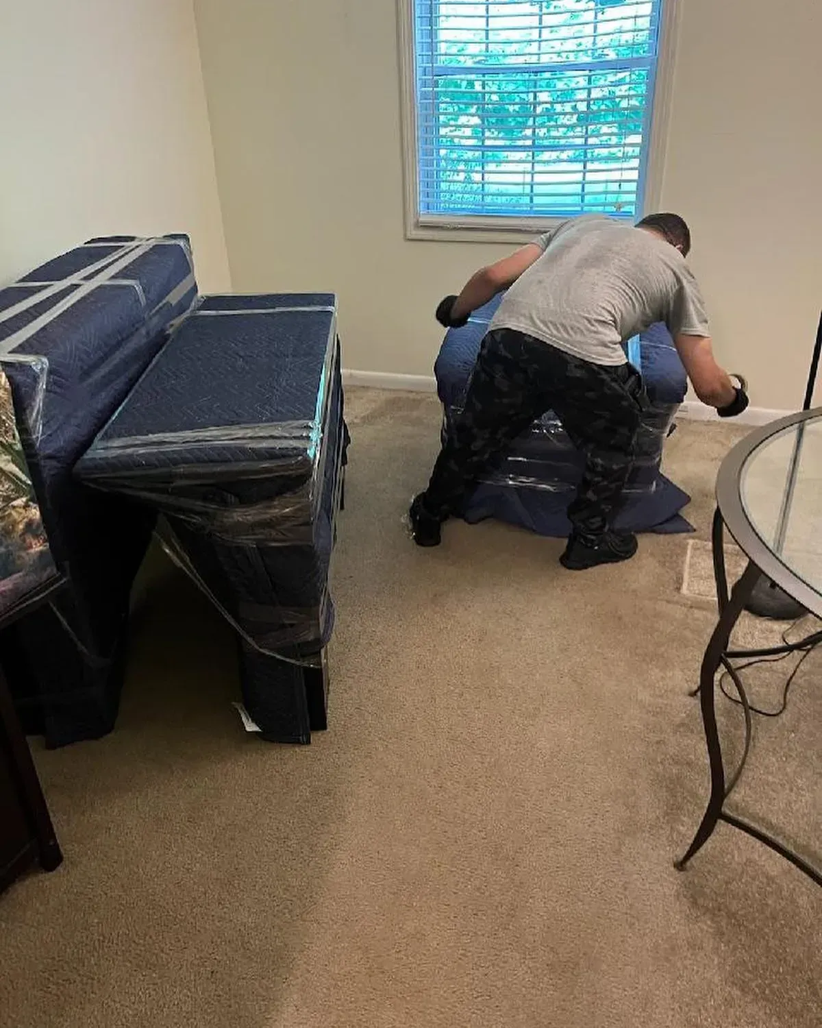 Person moving wrapped furniture in a room with a window, carpet, and dining table.