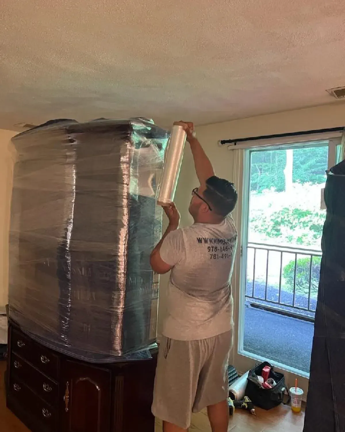 Person wrapping a large wooden cabinet in clear plastic, near a window.