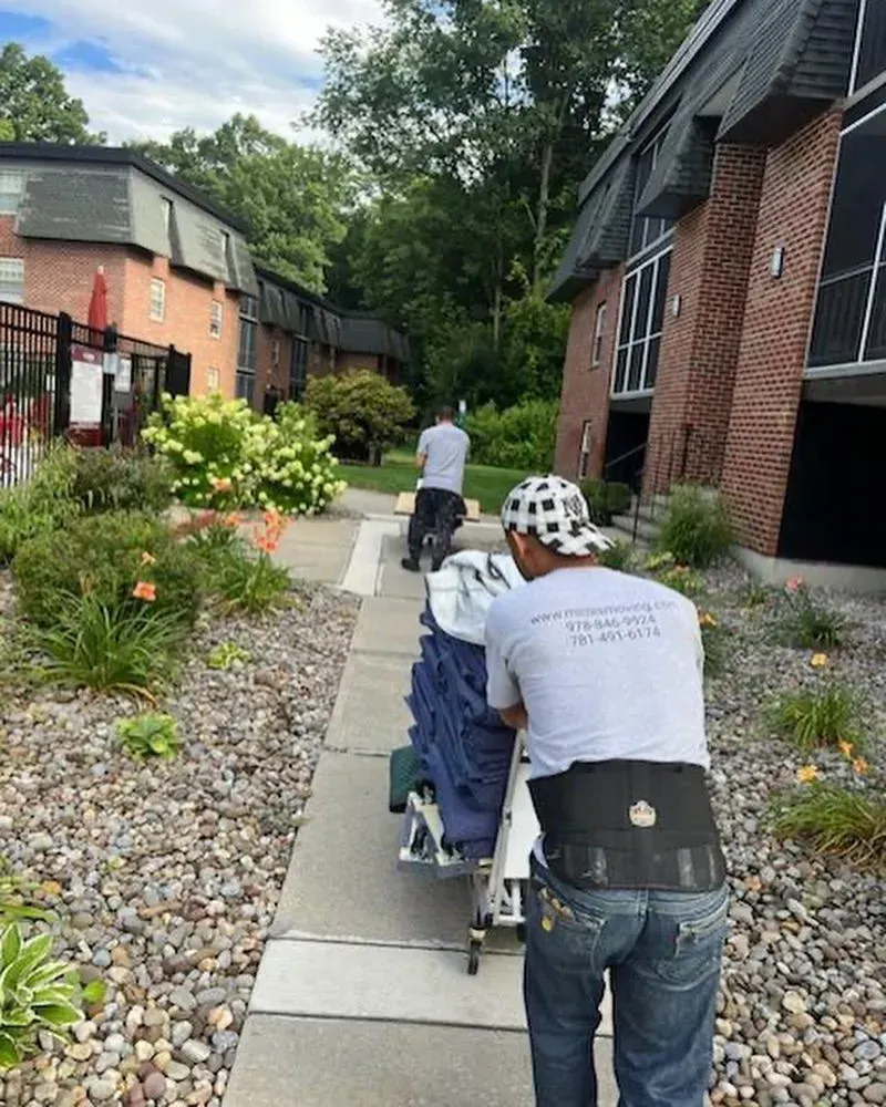 Two people move items on a dolly along a sidewalk between apartment buildings.