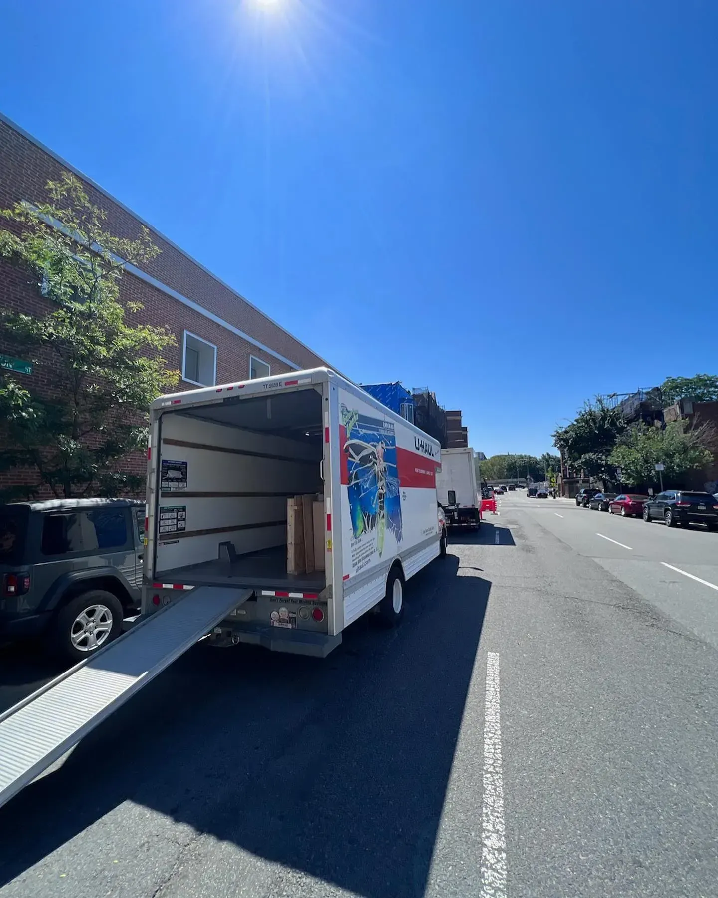 U-Haul truck parked on a street with its ramp down. Clear blue sky, sunny day. Another vehicle behind.