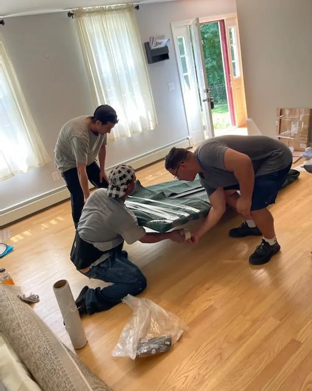Three people moving a green tarp-covered object inside a room with hardwood floors and bright windows.