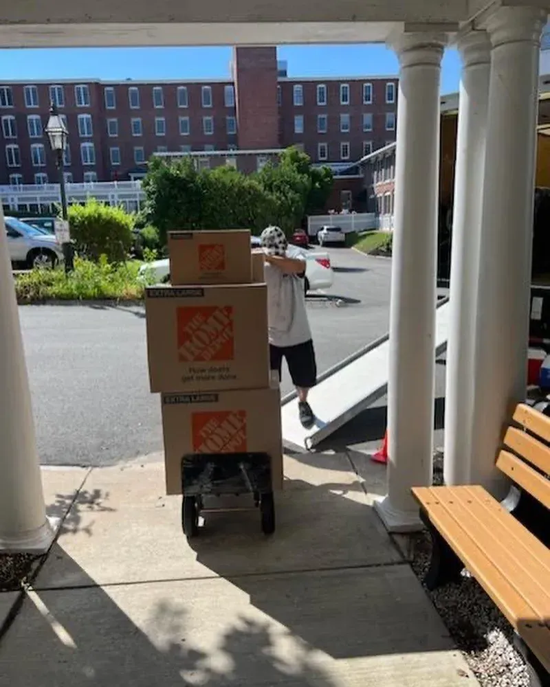 Person using a hand truck to move stacked moving boxes from a building entrance.