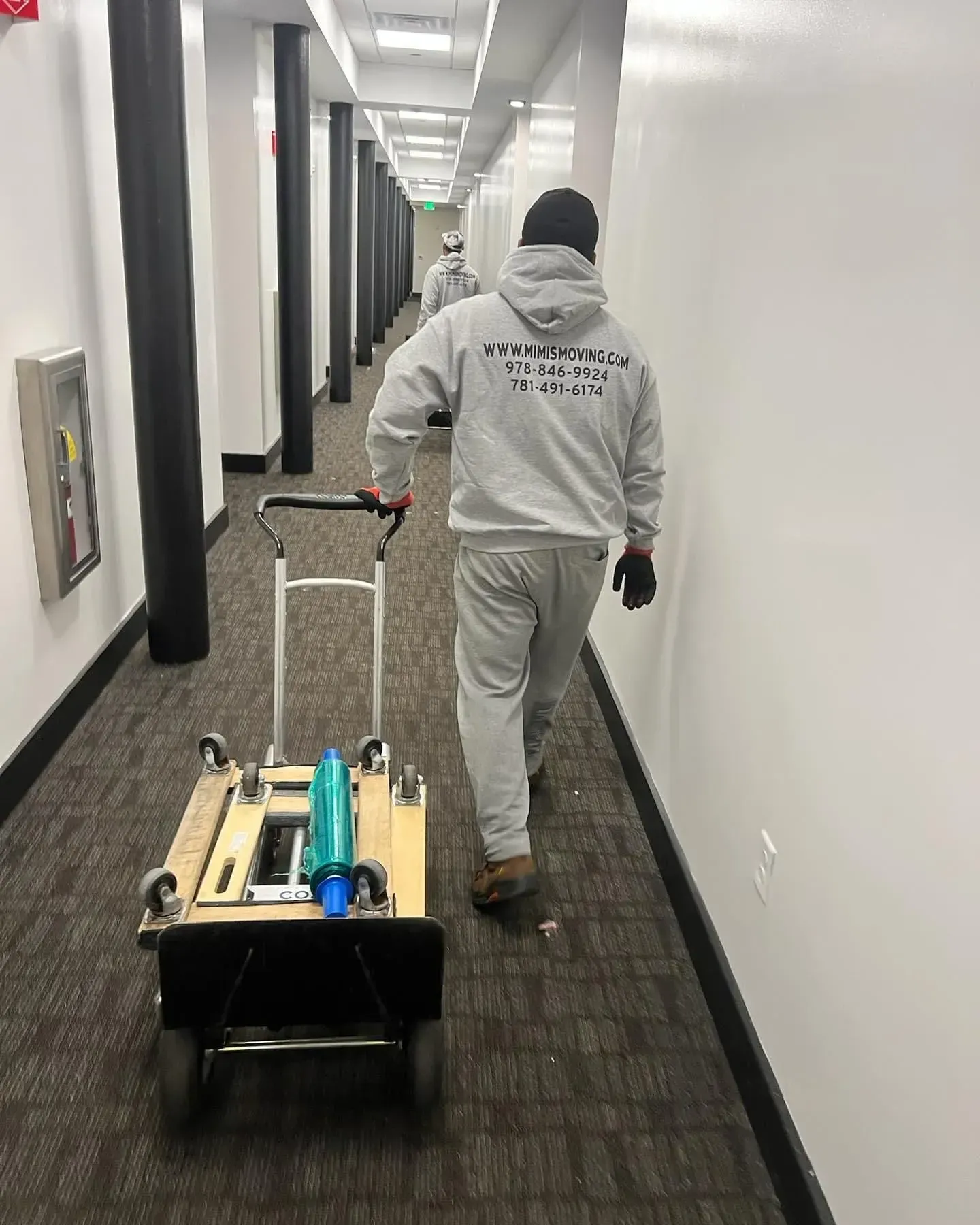 Person in gray sweatsuit pulling a cart with equipment down a long hallway with white walls and carpet.