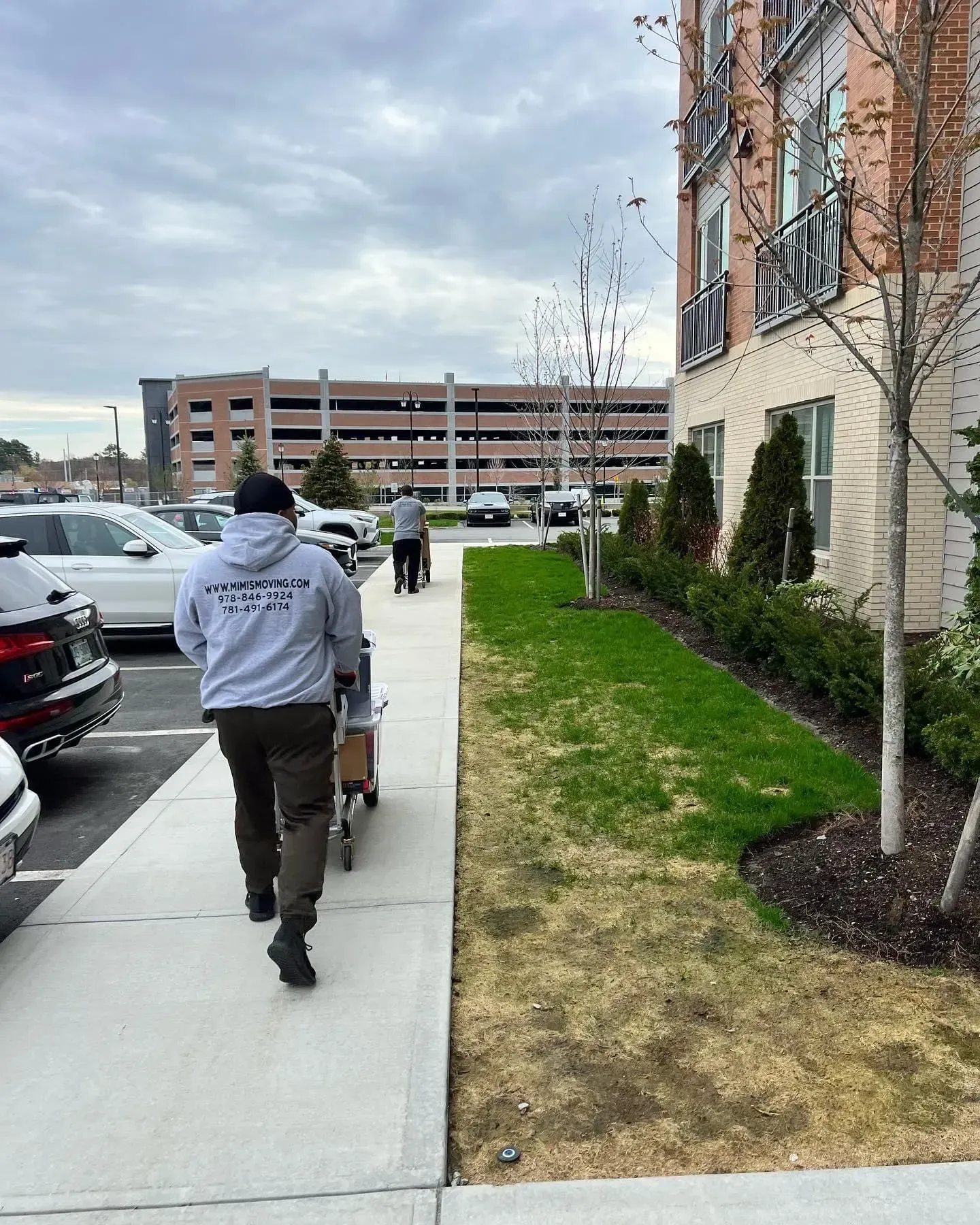 Person pulling a cart on a sidewalk next to a building and lawn. Cars and parking garage are in the background.