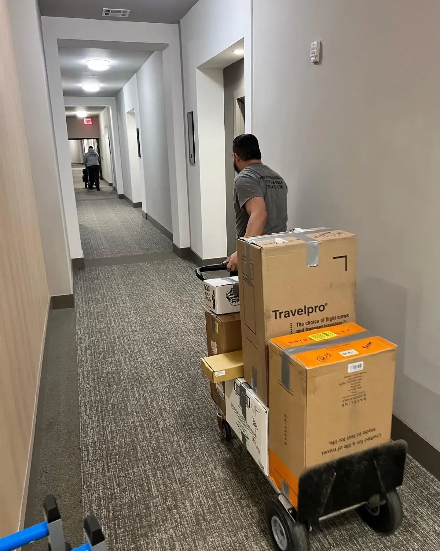 Man pushing a hand truck loaded with boxes down a long hallway with a second person at the end.